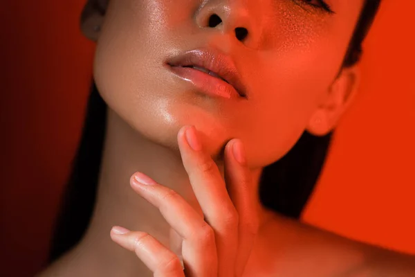 Vue recadrée de belle asiatique fille toucher brillant visage dans rouge éclairage — Photo de stock