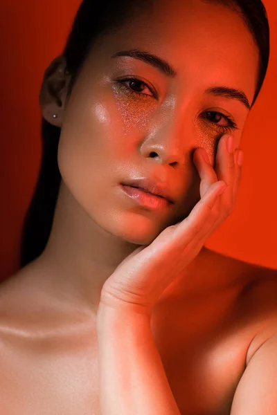 Hermosa chica asiática desnuda tocando la cara con brillantes de plata en la cara y los ojos cerrados en la iluminación roja - foto de stock