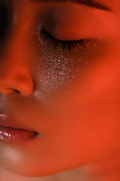 Close up view of beautiful asian girl with silver sparkles on face and closed eyes in red lighting — Stock Photo