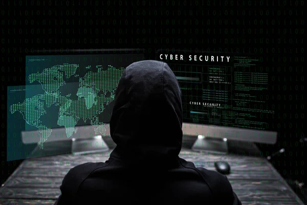 Vista posterior del hacker encapuchado sentado cerca de monitores de computadora con letras de seguridad cibernética en negro - foto de stock