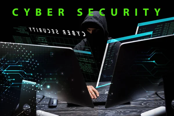 Hacker in hood sitting near computer monitors near cyber security lettering on black — Stock Photo