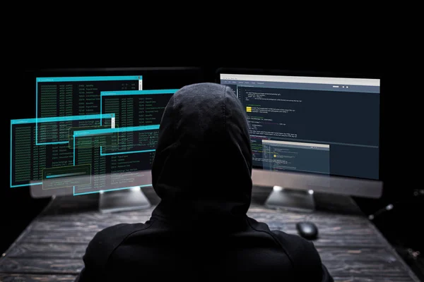 Back view of hooded hacker sitting near computer monitors with data on screens on black — Stock Photo