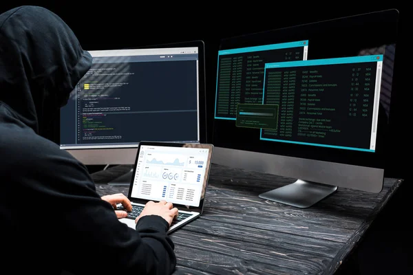 Hacker utilizando el ordenador portátil con gráficos y gráficos en la pantalla cerca de monitores de ordenador en negro - foto de stock