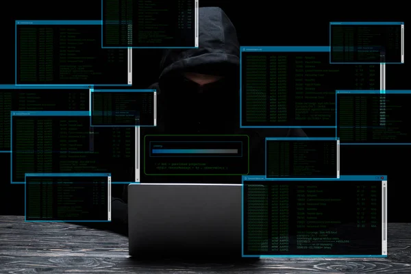 Hacker in hood using laptop near screens with data on black — Stock Photo