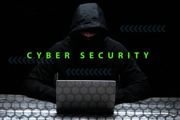 Hacker en el capó usando el ordenador portátil cerca de letras de seguridad cibernética en negro - foto de stock