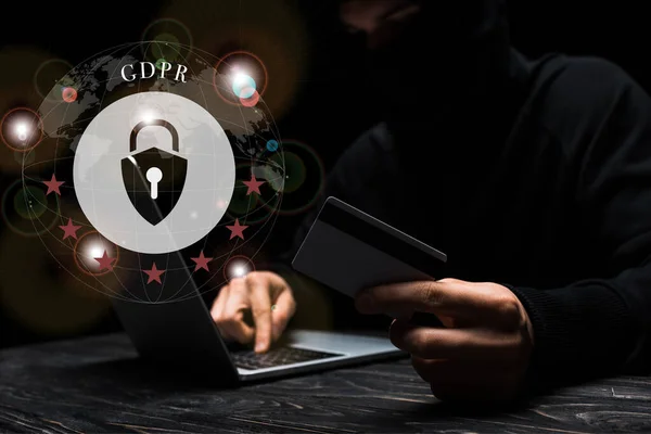 Selektiver Fokus des Hackers in Maske mit Laptop, während er Kreditkarte in der Nähe eines Vorhängeschlosses mit gdpr-Schriftzug auf schwarz hält — Stockfoto