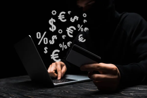 Selective focus of hacker in mask using laptop while holding credit card near money signs on black — Stock Photo