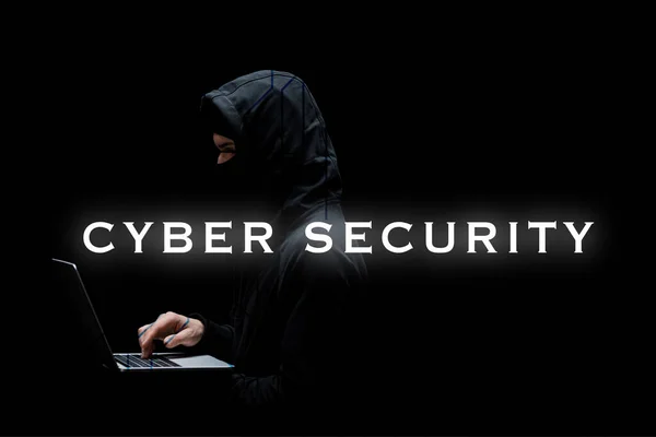 Anonymous hacker in hood and mask using laptop near cyber security lettering on black — Stock Photo