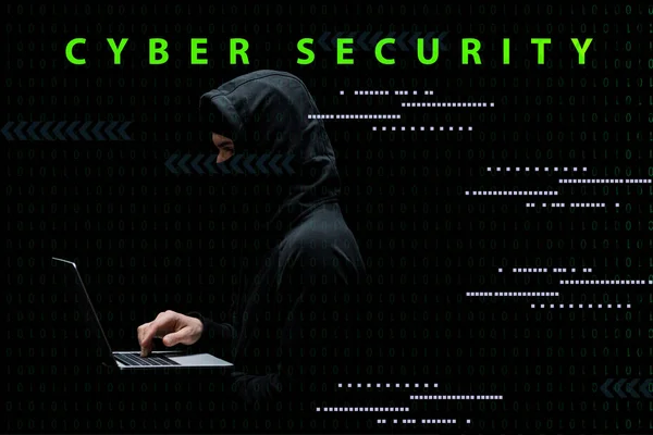 Anonimo uomo in cappuccio e maschera utilizzando laptop vicino alla cibersicurezza lettering su nero — Foto stock