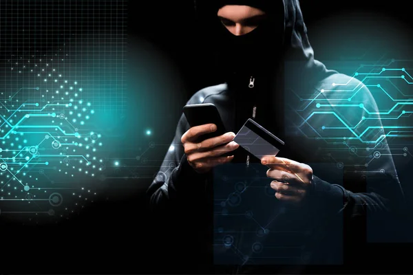 Hacker in hood using smartphone and holding credit card near illustration on black — Stock Photo