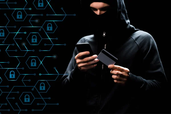Hacker in hood using smartphone and holding credit card near padlocks on black — Stock Photo