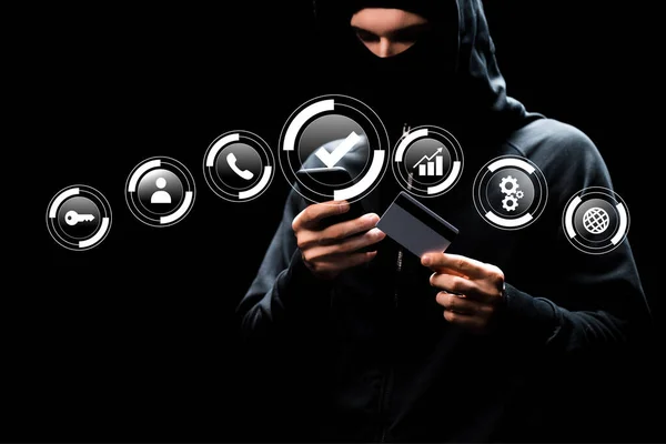 Hacker in hood using smartphone and holding credit card near web icons on black — Stock Photo