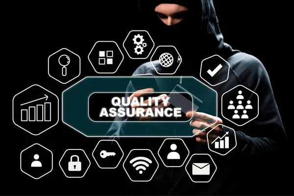 Hacker in hood using smartphone and holding credit card near quality assurance lettering on black — Stock Photo