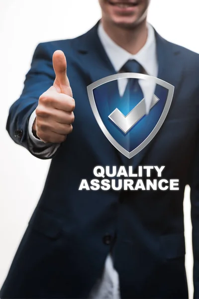 Selective focus of happy businessman in suit showing thumb up near quality assurance lettering on white — Stock Photo