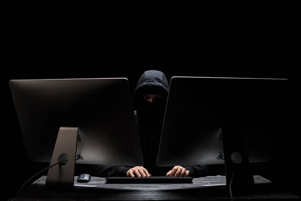 З капюшоном хакер в масці, що сидить біля комп'ютерних моніторів ізольовані на чорному — стокове фото
