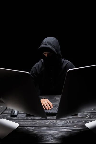 Человек в капюшоне в маске сидит рядом с мониторами компьютера, изолированными на черном — стоковое фото