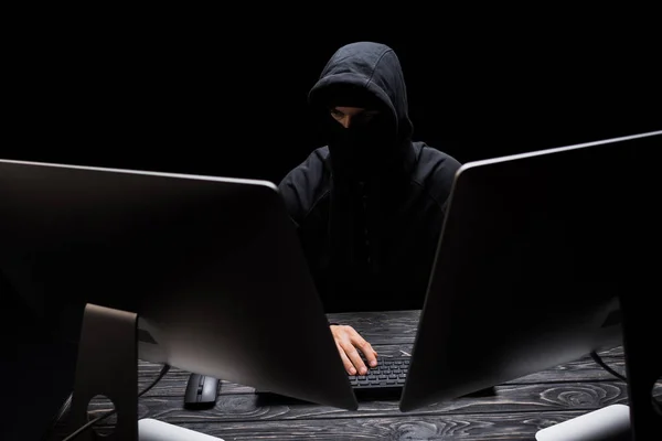 Hacker em máscara sentado perto de monitores de computador isolado em preto — Fotografia de Stock