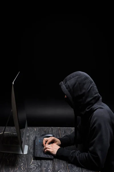 Hacker incappucciato in maschera digitando sulla tastiera del computer su nero — Foto stock