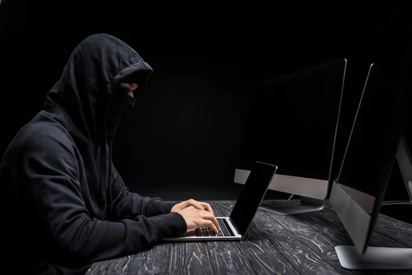 Back view of hooded hacker with hands behind head near computer monitors with blank screen isolated on black — Stock Photo