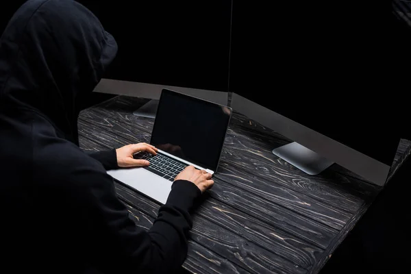 Hooded hacker using laptop near computer monitors with blank screen isolated on black — Stock Photo