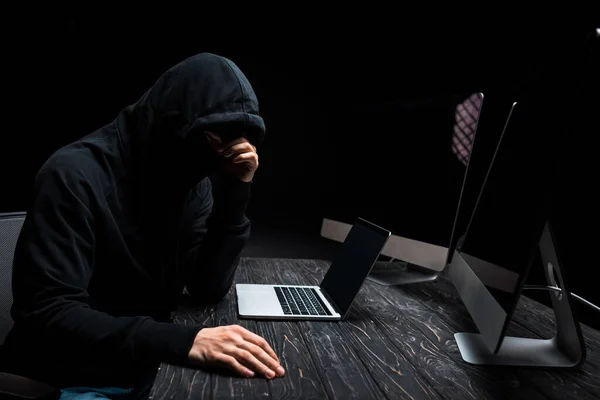 Upset hacker sitting near laptop and computer monitors with blank screen isolated on black — Stock Photo