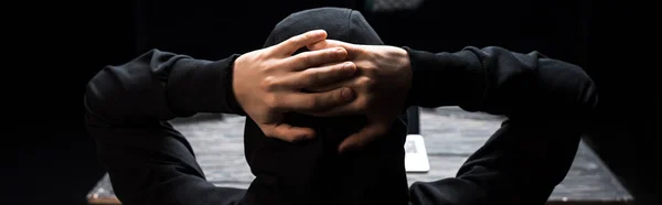 Panoramic shot of hooded hacker with hands behind head isolated on black — Stock Photo