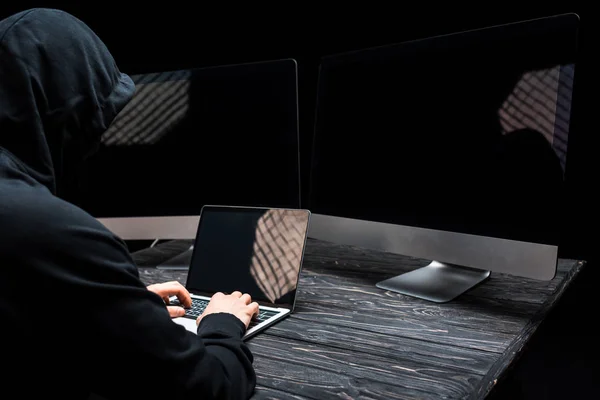 Hacker using laptop with blank screen near computer monitors isolated on black — Stock Photo