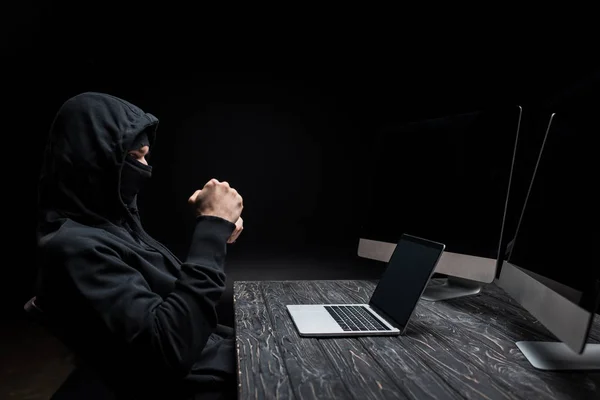 Hacker con los puños cerrados mirando a la computadora portátil con pantalla en blanco cerca de monitores de ordenador aislados en negro - foto de stock