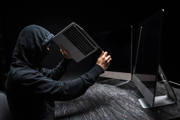 Hacker holding laptop near computer monitors with blank screen isolated on black — Stock Photo