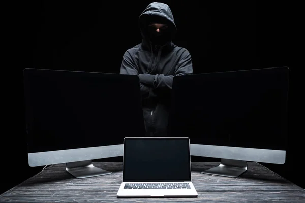 Hacker in mask standing with crossed arms near computer monitors and laptop with blank screen  isolated on black — Stock Photo