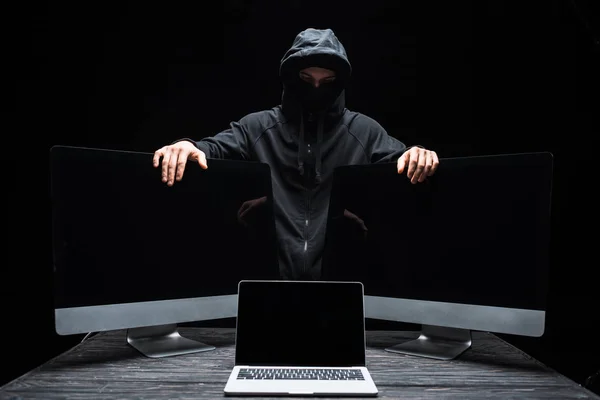 Hacker en la máscara tocando monitores de ordenador cerca de la computadora portátil con pantalla en blanco aislado en negro - foto de stock