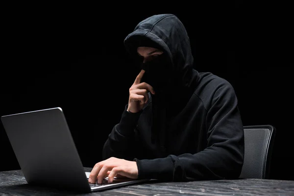 Young hacker touching mask and using laptop isolated on black — Stock Photo