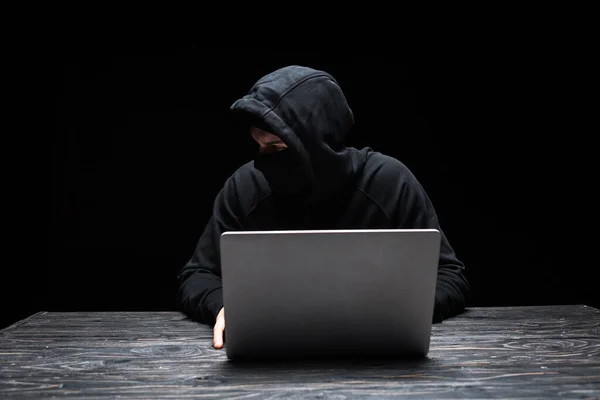 Young hacker in mask looking away while using laptop isolated on black — Stock Photo