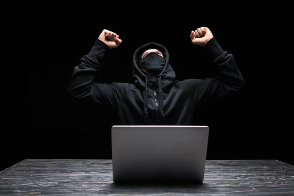 Excited hacker in mask celebrating triumph near laptop isolated on black — Stock Photo