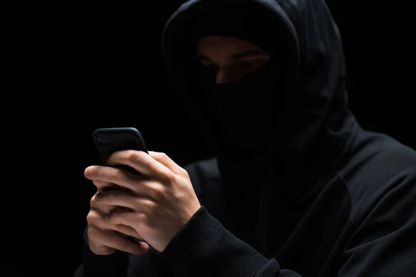 Selective focus of hacker in mask using smartphone isolated on black — Stock Photo