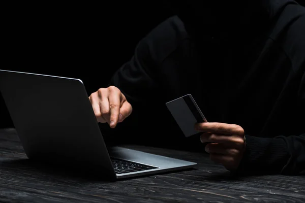 Cropped view of hacker using laptop while holding credit card isolated on black — Stock Photo