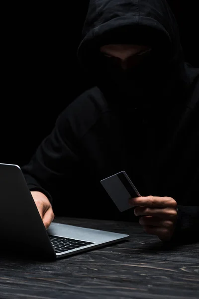 Pirate dans le capot et le masque à l'aide d'un ordinateur portable tout en tenant la carte de crédit isolé sur noir — Photo de stock