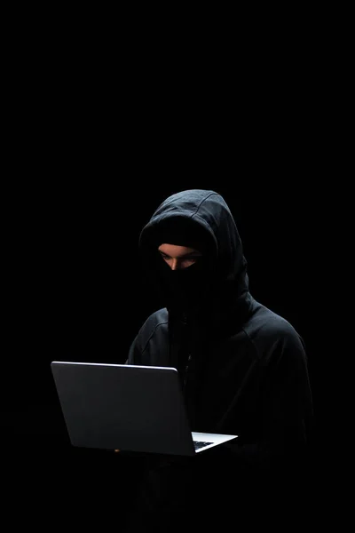 Young hacker in hood using laptop isolated on black — Stock Photo