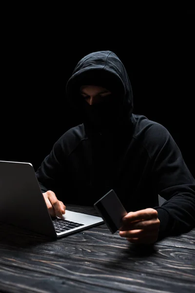 Hacker no capuz usando laptop enquanto segurando cartão de crédito isolado no preto — Fotografia de Stock