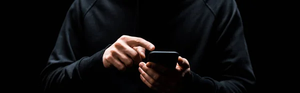 Panoramic shot of hacker pointing with finger at smartphone isolated on black — Stock Photo