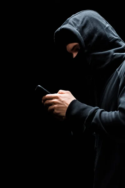 Hacker anonimo in cappuccio e maschera utilizzando smartphone isolato su nero — Foto stock