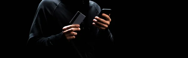 Panoramic shot of young hacker using smartphone and holding credit card isolated on black — Stock Photo