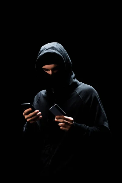 Jovem hacker no capuz e máscara usando smartphone e segurando cartão de crédito isolado no preto — Fotografia de Stock