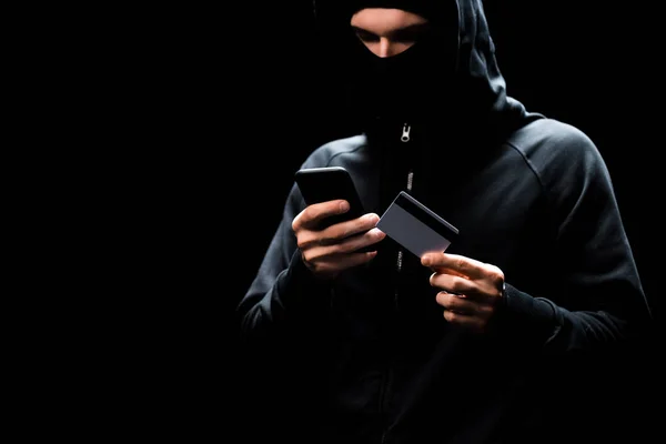 Hacker in hood using smartphone and holding credit card isolated on black — Stock Photo