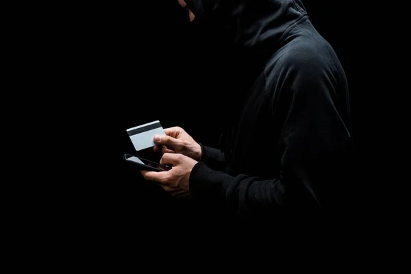 Cropped view of hacker in hood and mask using smartphone and holding credit card isolated on black — Stock Photo