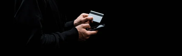 Panoramic shot of hacker using smartphone and holding credit card isolated on black — Stock Photo