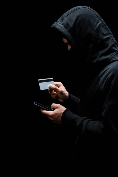 Hacker in hood and mask using smartphone and holding credit card isolated on black — Stock Photo