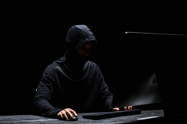 Pirate anonyme à capuchon dans le masque à l'aide d'un ordinateur isolé sur noir — Photo de stock
