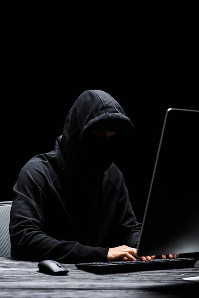 Hacker anônimo encapuzado na máscara digitando no teclado do computador isolado no preto — Fotografia de Stock