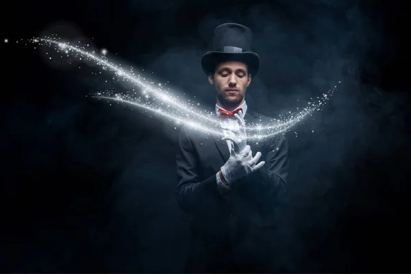 Magician in suit and hat wearing gloves in dark smoky room with glowing illustration — Stock Photo